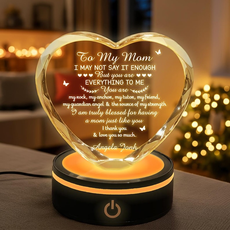 Puede incluir: Adorno de cristal en forma de coraz&oacute;n con el texto "To My Mom" y un mensaje de amor. El cristal transparente se asienta sobre una base negra con un brillo naranja y un bot&oacute;n de encendido. Un regalo reflexivo.