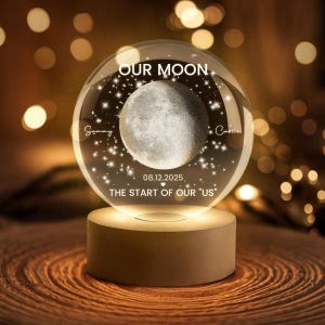 May include: A clear glass sphere with a detailed moon design, illuminated by a warm light. The sphere has the text "OUR MOON," names, and the date "08.12.2025," with the phrase "THE START OF OUR US." It sits on a wooden base.