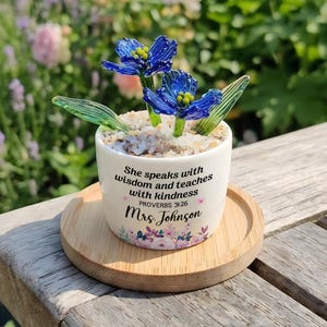 May include: A white ceramic planter on a round bamboo tray containing two blue glass flowers with green glass leaves and small pebbles. The planter features the text: She speaks with wisdom and teaches with kindness PROVERBS 31:26 Mrs. Johnson.