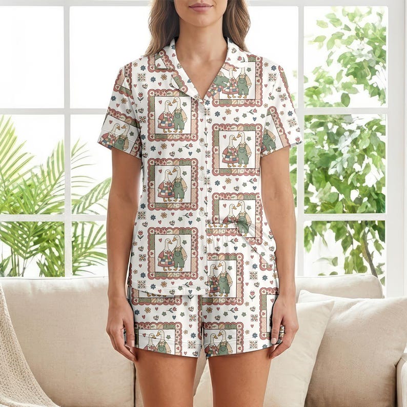 May include: White pajama set with a short-sleeved button-down shirt and matching shorts. The set features a repeating pattern of framed goose illustrations in a country style, with floral and heart accents. The pajama set is made of a soft, lightweight fabric.