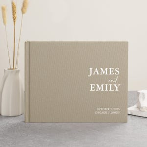 May include: A beige linen-covered wedding guest book with the names "JAMES and EMILY" in white script. The date "OCTOBER 3, 2025" and location "CHICAGO, ILLINOIS" are printed below the names. A white vase with wheat stalks is on the left.