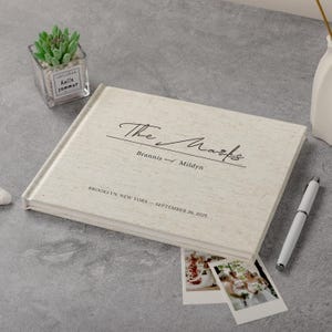 May include: A cream linen wedding album with the inscription "The Marks" and wedding details. A pen, two polaroid photos, and a small succulent plant in a glass container are also present.