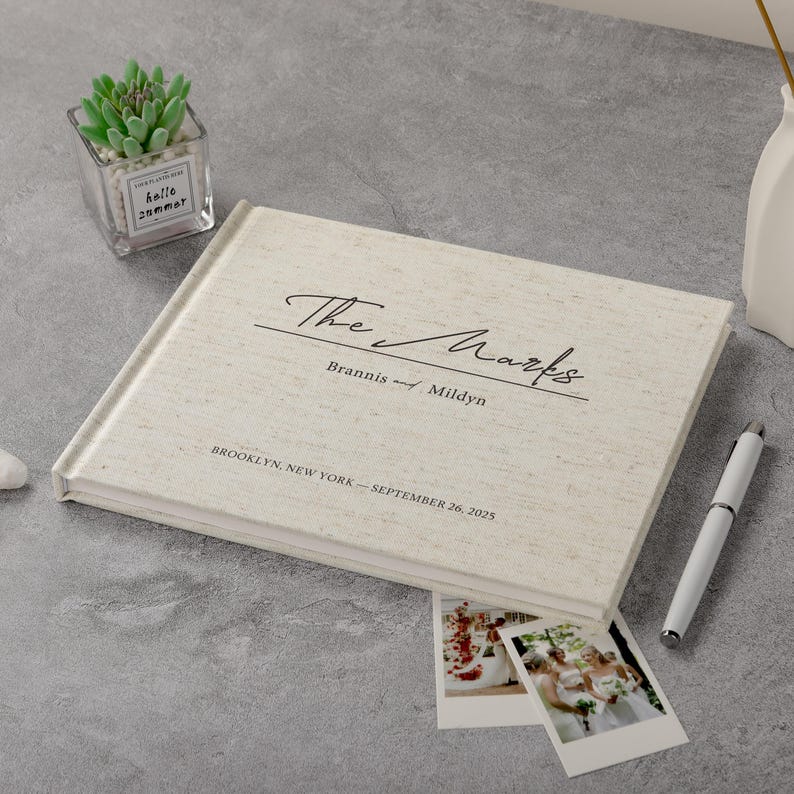 May include: A cream linen wedding album with the inscription "The Marks" and wedding details. A pen, two polaroid photos, and a small succulent plant in a glass container are also present.