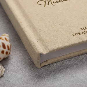 May include: A beige linen-covered book with the name "Michael" in cursive script. The book's corner is visible, showing the thickness of the pages. The text "MA LOS AN" is printed on the cover. A seashell is in the foreground.