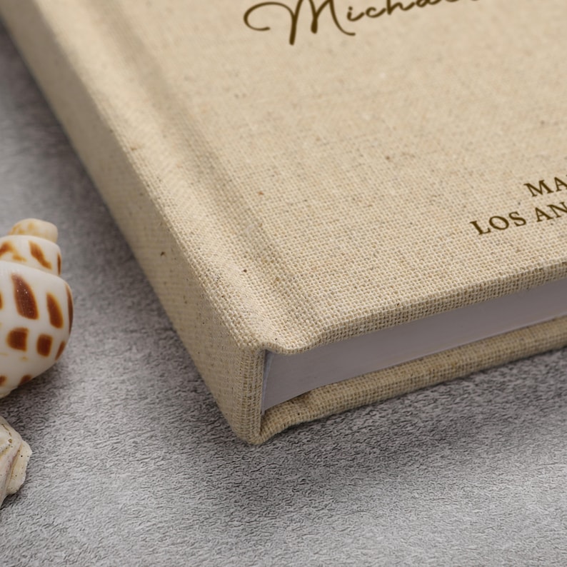 May include: A beige linen-covered book with the name "Michael" in cursive script. The book's corner is visible, showing the thickness of the pages. The text "MA LOS AN" is printed on the cover. A seashell is in the foreground.