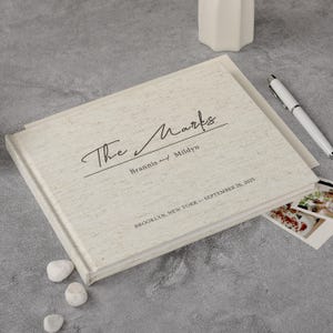 May include: A cream-coloured linen-textured photo album with the text "The Marks" in script. The album includes the names "Brannis and Mildyn", the date "September 26, 2025" and the location "Brooklyn, New York".