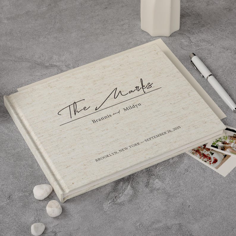 May include: A cream-coloured linen-textured photo album with the text "The Marks" in script. The album includes the names "Brannis and Mildyn", the date "September 26, 2025" and the location "Brooklyn, New York".