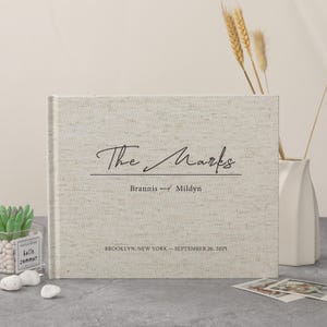 May include: A beige linen-covered photo album with the title "The Marks" in cursive script. The names "Brannis and Mildyn" and the date "September 26, 2025" are printed below, along with the location "Brooklyn, New York".