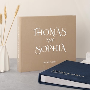 Peut inclure: Un livre d'or de mariage beige avec les noms "THOMAS AND SOPHIA" en écriture blanche et la date "07.JULY.2026". Un plus petit livre bleu marine avec "MICHAEL & ISABELLA WEDDING GUESTBOOK" est au premier plan. Vase décoratif avec des épis de blé.