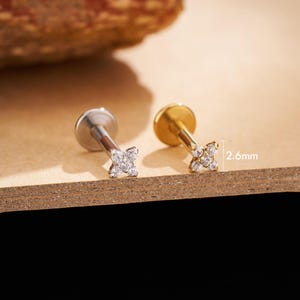 Può includere: Due piccoli orecchini a lobo con un design a fiore a quattro petali. Un orecchino è argentato, l'altro è dorato. Ogni fiore è incastonato con piccole gemme trasparenti. Gli orecchini sono esposti su una superficie marrone.