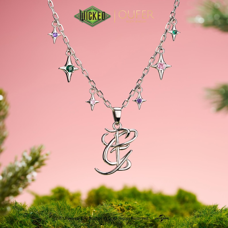 May include: A silver necklace with a pendant of stylized initials and star-shaped charms with coloured gemstones. The necklace has a delicate chain and is set against a pink background with the text "WICKED" and "QUEER".