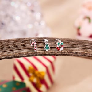 May include: Three Christmas stud earrings: a candy cane, a green Christmas tree, and a red and white wreath with a green bow. The earrings are silver-toned and set on a wooden surface. Festive holiday jewellery.