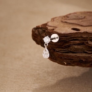 May include: A silver labret stud earring featuring a round, clear gemstone and a dangling pear-shaped gemstone. The earring is presented on a piece of wood against a light brown backdrop. This jewellery is designed for body piercings.
