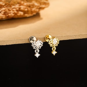 May include: Two small, decorative earrings. One is silver-toned, the other gold-toned. Each earring features a lotus flower design with a dangling, small, clear gemstone. The earrings are set against a neutral background.