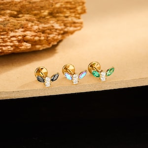 May include: Three gold-colored stud earrings with leaf-like designs. Each earring features a cluster of gemstones: black and white, iridescent and white, and green and white. The earrings are displayed on a light brown surface.