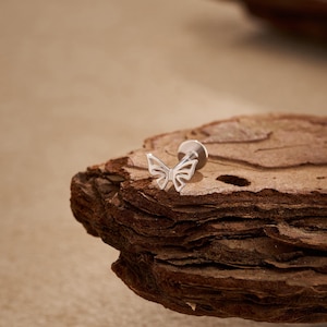 May include: A silver butterfly-shaped earring stud. The earring is set against a textured, brown wooden surface. The butterfly design is delicate and detailed, with open wings.