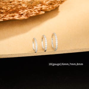 Może przedstawiać: Trzy srebrne kolczyki koła z małymi, przezroczystymi kamieniami są prezentowane na jasnobrązowej powierzchni. Kolczyki są w różnych rozmiarach. Tekst na dole brzmi "18(gauge)/6mm,7mm,8mm".