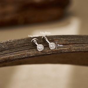 May include: Two silver-colored, flat-back earrings with clear, round gemstones. The earrings are displayed on a wooden surface. Measurements are provided: 3mm, 16g, 6.6mm, and 16g, 8mm. The earrings are designed for body piercings.