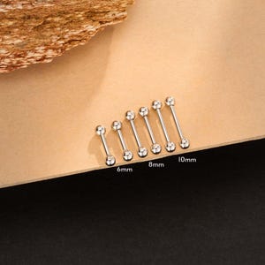 Puede incluir: Una colección de pendientes de barra de plata en varios tamaños, incluyendo 6 mm, 8 mm y 10 mm. Los pendientes tienen un acabado pulido y una bola en cada extremo. Se muestran sobre una superficie marrón claro.