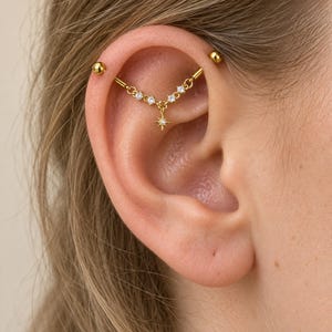 May include: A gold-toned industrial barbell earring featuring a central chain segment adorned with four small clear round stones and a dangling star-shaped charm with a single clear stone at its center, secured with two smooth spherical end balls.
