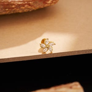 May include: A gold-colored flower-shaped stud earring with six clear, marquise-cut cubic zirconia stones. The earring is set on a gold post and backing. The jewelry is displayed on a light brown surface.