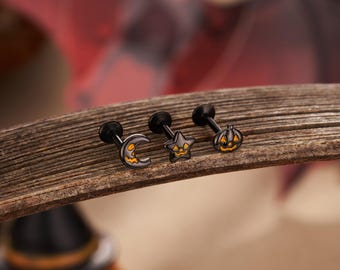 Op de afbeelding: Drie zwarte lip piercing studs met Halloween-thema. De studs hebben een halve maan, een ster en een pompoenontwerp. Elke stud heeft een zwarte paal en een platte, decoratieve bovenkant met oranje en gele accenten.