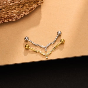 Op de afbeelding: Twee barbell-lichaamssieraden, één in zilverkleur en één in goudkleur. Elk sieraad heeft een V-vormige hanger versierd met kleine, heldere, marquise-geslepen stenen en een enkele ronde steen in het midden, bevestigd aan een rechte staaf.