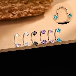 May include: A selection of silver nose rings, each with a curved design and two gemstones. The gemstones are in various colours, including iridescent, clear, black, purple, pink, and turquoise. The jewellery is displayed on a brown surface.