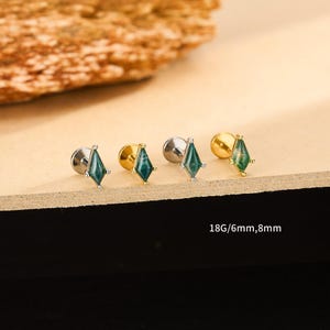 Puede incluir: Cuatro pendientes de botón con una piedra en forma de diamante de color verde azulado. Dos pendientes tienen un poste plateado y dos tienen un poste dorado. El texto "18G/6mm,8mm" es visible.
