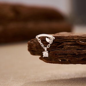 May include: A silver earring with a small chain and a dangling, round, clear gemstone. The earring is set against a textured, brown wooden surface, with a blurred background.