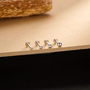 Può includere: Quattro orecchini a bottone color argento con sfere di diverse dimensioni sono presentati su una superficie di legno. Gli orecchini hanno un retro piatto e un perno. Gli orecchini sono disposti in fila, mostrando le loro variazioni di dimensioni.