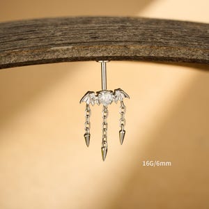 Può includere: Un piercing per ombelico color argento con un design ad ali di pipistrello e catene pendenti. La pietra centrale è trasparente e a forma di cuore. Le catene terminano con forme a cono appuntito. Il testo "16G/6mm" è visibile.