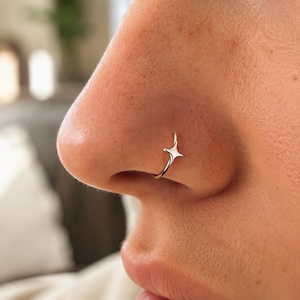 May include: A polished silver-toned nose ring featuring a small, four-pointed star charm. The thin, circular hoop is designed to fit snugly around the nostril, with the star element positioned prominently on the outer edge of the nose piercing.