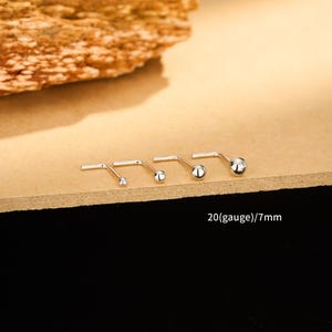 May include: Four silver nose studs with ball ends, displayed on a light brown surface. The text "20(gauge)/7mm" is visible. The studs are arranged in a row, showcasing different sizes.