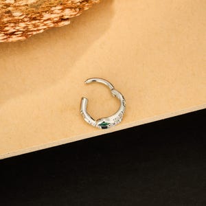 May include: A silver-colored hinged hoop earring with a small green gem and several clear gems. The earring is open, showing the clasp. The earring is on a light brown surface.