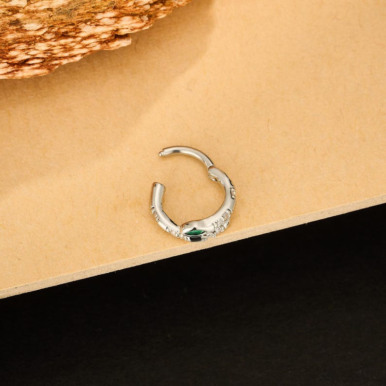 May include: A silver-colored hinged hoop earring with a small green gem and several clear gems. The earring is open, showing the clasp. The earring is on a light brown surface.