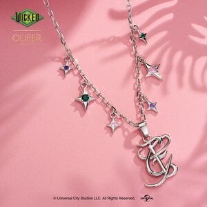 May include: A silver necklace featuring a chain and star-shaped charms, each with a different coloured gem. The necklace has a pendant with the letters "FY" in a stylised font. The background is pink, and the image includes the text "WICKED" and "OUFER".