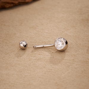 Puede incluir: Una joya de piercing plateada tipo barra con una gema redonda y transparente engastada en un bisel. También se ve una pequeña bola plateada separada. La joya está diseñada para piercing.