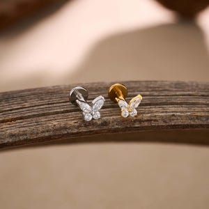 May include: Two butterfly-shaped earrings, one silver-toned and one gold-toned, each adorned with clear, sparkling gemstones. The earrings are displayed on a textured, wooden surface, showcasing their delicate design and intricate details. The butterfly earrings are a jewelry item.