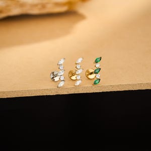 May include: Three ear studs with a curved design. One stud is silver with white gems, the second is gold with white gems, and the third is gold with green gems. The studs are displayed on a light brown surface.