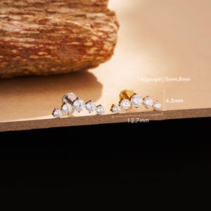 May include: Two ear studs, one silver and one gold, each featuring a curved row of clear, sparkling gemstones. The silver stud is on the left, and the gold stud is on the right. The studs are 12.7mm long and 4.5mm high.