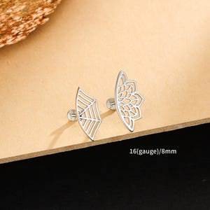 Può includere: Due orecchini a lobo color argento. Uno presenta un design a ragnatela, l'altro un motivo floreale. Il testo "16(gauge)/8mm" è visibile. Gli orecchini sono esposti su una superficie marrone chiaro.