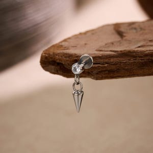 Può includere: Un piercing per il corpo color argento con una gemma rotonda e trasparente e un ciondolo a forma di cono pendente. Il gioiello è esposto su uno sfondo marrone strutturato.