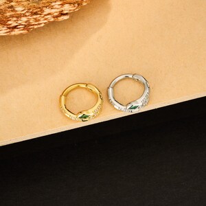May include: Two hoop earrings, one gold-toned and one silver-toned, each featuring a snake design. The snakes have green eyes and are adorned with small, clear gemstones. The earrings are displayed on a light brown surface.