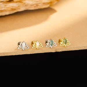May include: Four gold and silver lotus flower stud earrings with white and green accents. The earrings are displayed on a light brown surface with a dark brown background. The lotus flower design is detailed and elegant.
