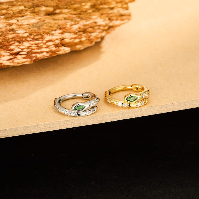 May include: Two snake-shaped hoop earrings. One is silver-toned, the other gold-toned. Both have a green gemstone for the snake's eye and are encrusted with small, clear gemstones. The earrings are displayed on a light brown surface.