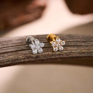 May include: Two flower-shaped ear studs. One is silver-toned, the other gold-toned. Each stud features a cluster of clear, round gemstones arranged in a floral pattern. The studs are displayed on a piece of weathered wood, showcasing jewelry.