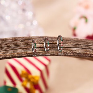 May include: Three silver hoop earrings with red, green, and white gemstones. The earrings are 6mm, 7mm, and 8mm in diameter. They are displayed on a wooden surface with a blurred background.