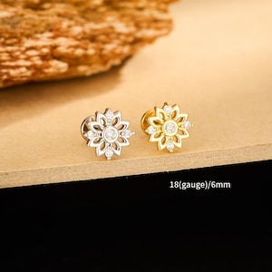 Może przedstawiać: Dwa kolczyki w kształcie kwiatów, jeden srebrny i jeden złoty, każdy z centralnym kamieniem i mniejszymi kamieniami. Kolczyki mają średnicę 6 mm.