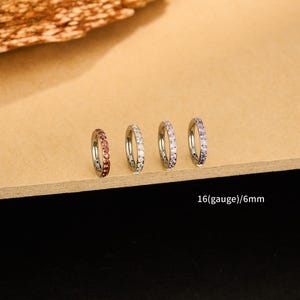 Puede incluir: Cuatro pendientes de aro plateados con piedras preciosas de diferentes colores: rojo, blanco y morado. El texto de la imagen dice "16(gauge)/6mm". Cada pendiente tiene 6 mm de diámetro.